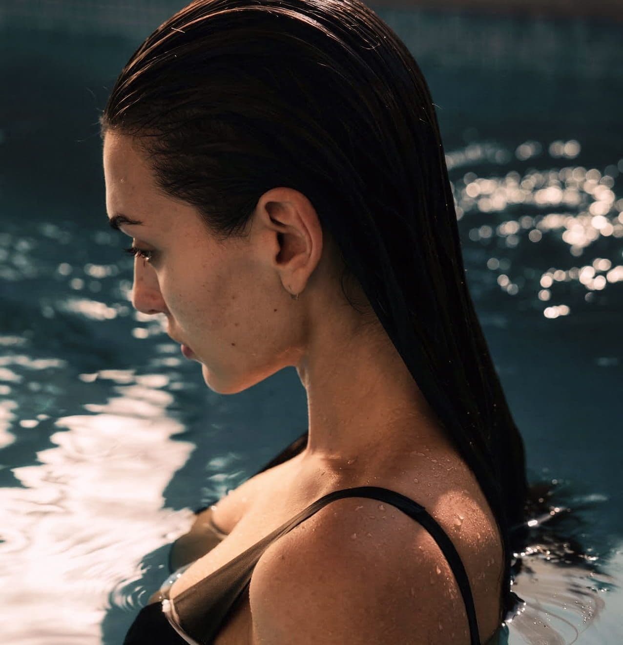 Woman with long dark hair emerging from sparkling blue pool water.